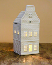 Load image into Gallery viewer, White ceramic house-shaped lantern with lit interior on a textured surface.
